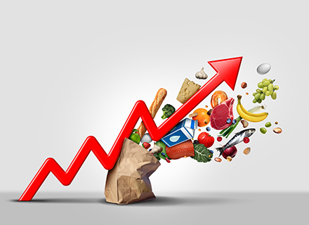 An upward-pointing red arrow emerges from a paper bag filled with various groceries, symbolizing the rising cost of goods and services, a visual representation of inflation.